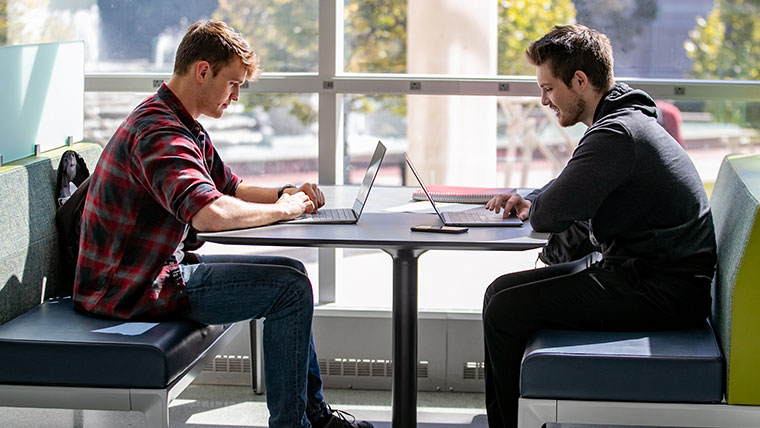Two Missouri State students facing each other at a table in the library with their laptops. 